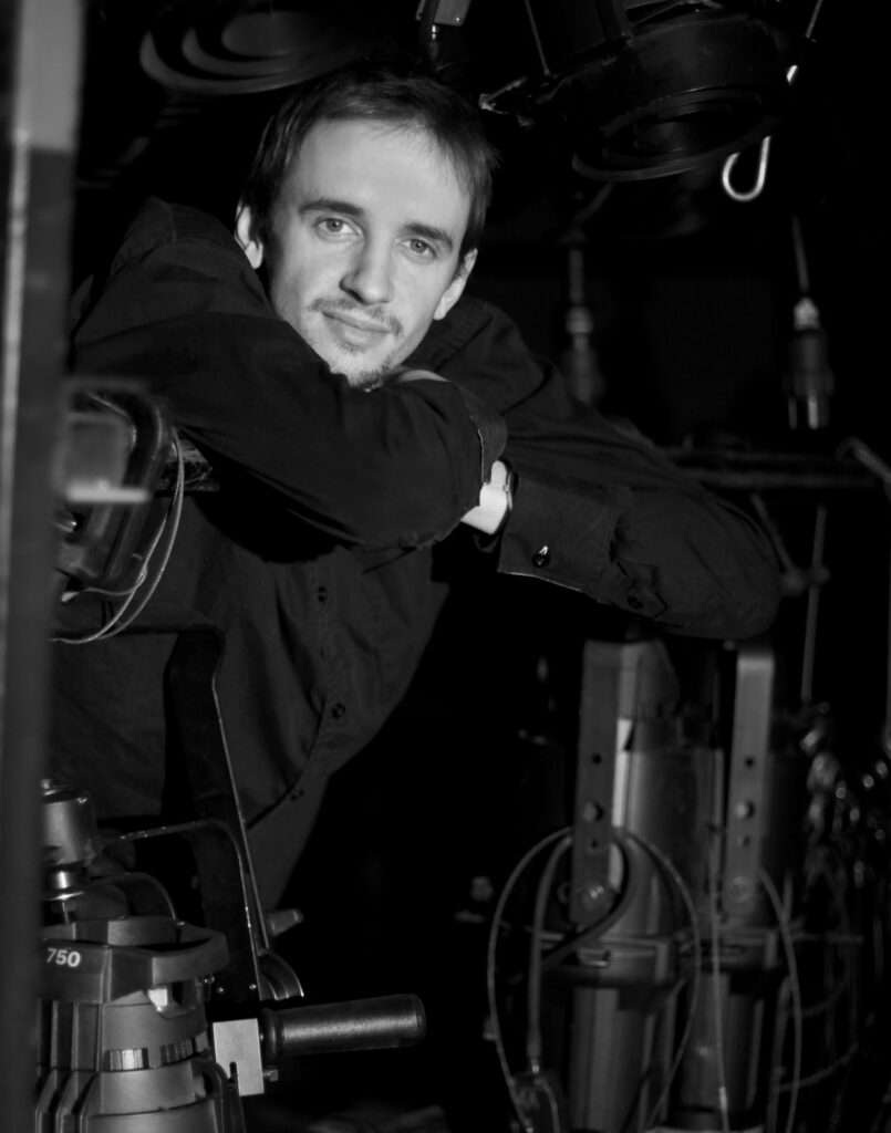 Black-and-white portrait of a smiling man leaning on film equipment, surrounded by studio lights and cables, creating a candid, friendly mood.